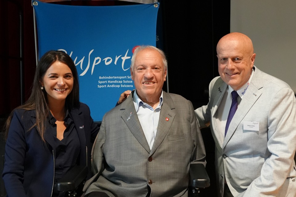 Drei Personen in Business-Kleidung stehen lächelnd vor einem blauen Banner mit der Aufschrift „PluSport“, einem Hinweis auf den Schweizer Behindertensport. Der Mann in der Mitte sitzt im Rollstuhl.