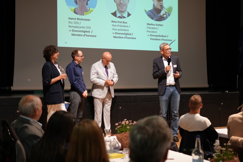 Vier Personen stehen auf der Bühne und sprechen bei einer Veranstaltung, während das Publikum zusieht. Eine Projektion im Hintergrund zeigt Namen und Fotos der Personen und suggeriert damit eine formelle Präsentation oder Preisverleihung.