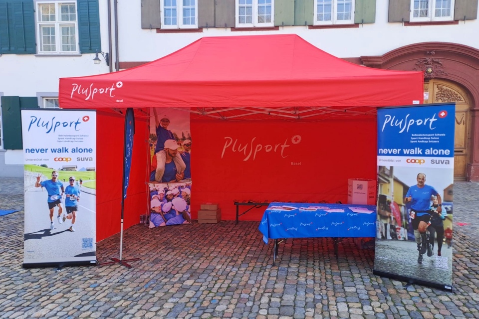 Ein rotes Plusport-Zelt ist auf dem Kopfsteinpflaster vor einem Gebäude aufgebaut. Zwei Banner zeigen Läufer mit dem Slogan "never walk alone". Unter dem Zelt stehen ein blauer Tisch und Werbematerialien.
