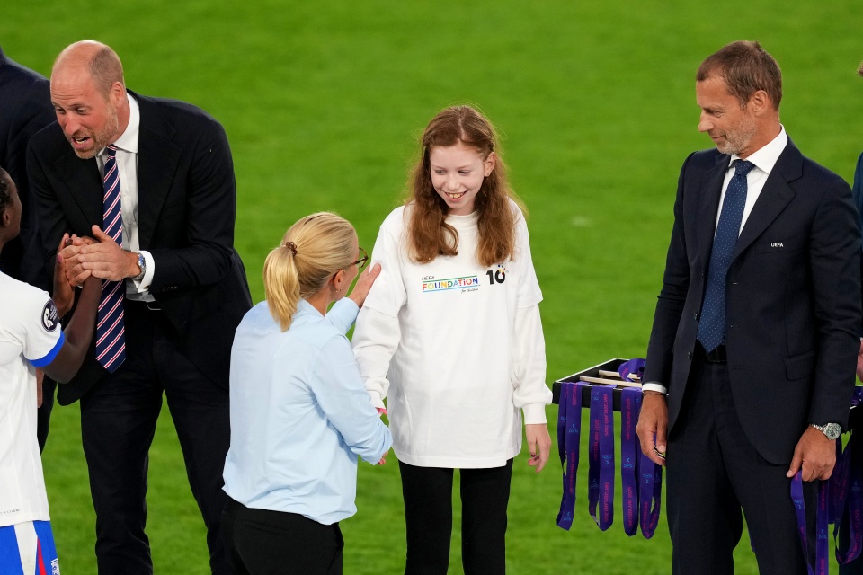 Ein junges Mädchen in einem weißen Hemd schüttelt lächelnd einer Frau in Blau die Hand, während zwei Männer in Anzügen zuschauen und Medaillen auf einem Tisch in der Nähe auf einem grünen Sportplatz ausgestellt sind.