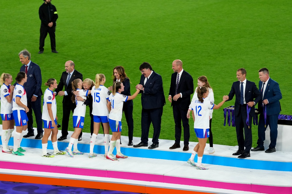 Fußballspieler in weißen Trikots stellen sich nach einem Spiel auf einem Podium auf, um von Offiziellen in Anzügen Medaillen entgegenzunehmen, mit grünem Rasen im Hintergrund.