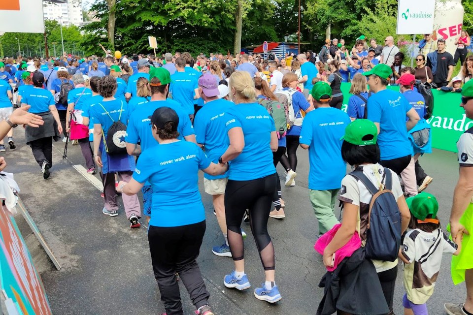 Eine große Gruppe von Menschen in passenden blauen Hemden und Sportkleidung läuft oder geht gemeinsam bei einer Outdoor-Veranstaltung. Einige Kinder tragen grüne Hüte, und Zuschauer stehen am Rand. Im Hintergrund sind Bäume und Veranstaltungsbanner zu sehen.