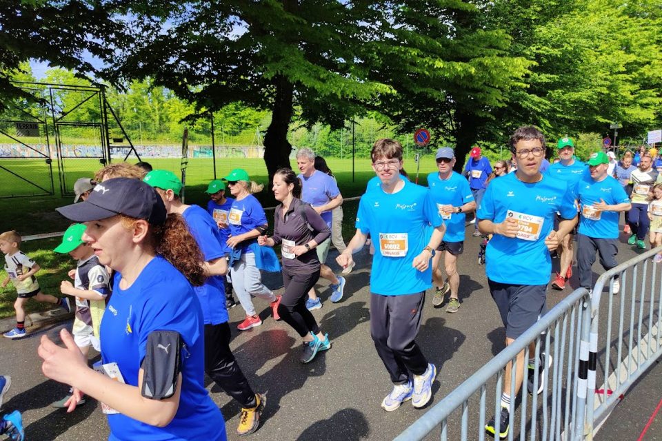 Eine Gruppe von Menschen in Sportkleidung und mit nummerierten Startnummern läuft an einem sonnigen Tag gemeinsam bei einem Rennen im Freien, umgeben von grünen Bäumen und Zuschauern hinter Absperrungen.