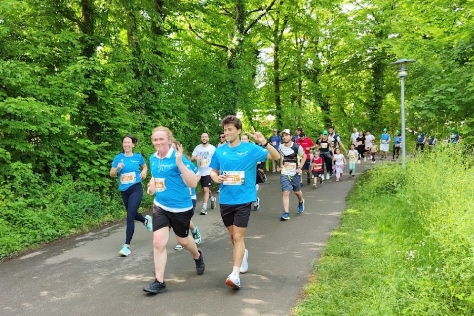 Eine Gruppe von Menschen läuft auf einem gepflasterten Weg, umgeben von üppigen grünen Bäumen. Zwei lächelnde Läufer in blauen Shirts führen die Menge an. Es scheint ein Gemeinschaftslauf oder eine Spaßveranstaltung an einem sonnigen Tag zu sein.
