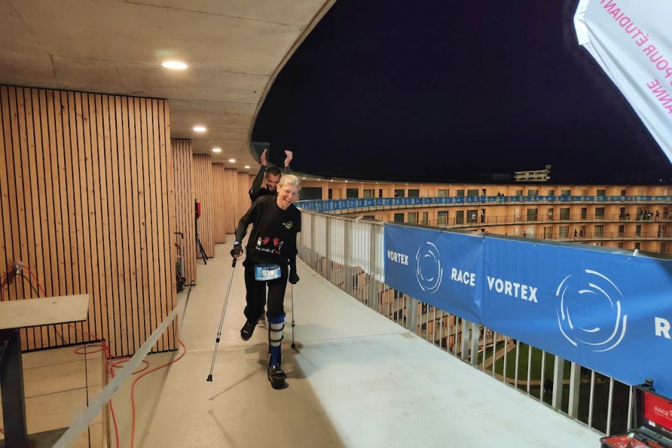 Ein lächelnder Sportler mit einer Beinprothese und Krücken läuft während einer nächtlichen Rennveranstaltung eine Rampe entlang, während blaue Race Vortex-Banner das Geländer säumen und eine weitere Person hinter ihm jubelt.