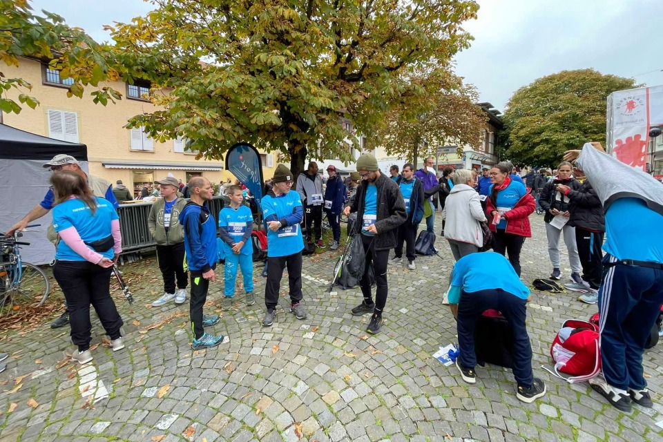 Eine Gruppe von Menschen, viele in blauen Hemden, versammelt sich auf einer Kopfsteinpflasterstraße in der Nähe von Bäumen und Gebäuden, möglicherweise vor oder nach einer Rennveranstaltung. Einige unterhalten sich, während andere ihre Taschen und Habseligkeiten sortieren.