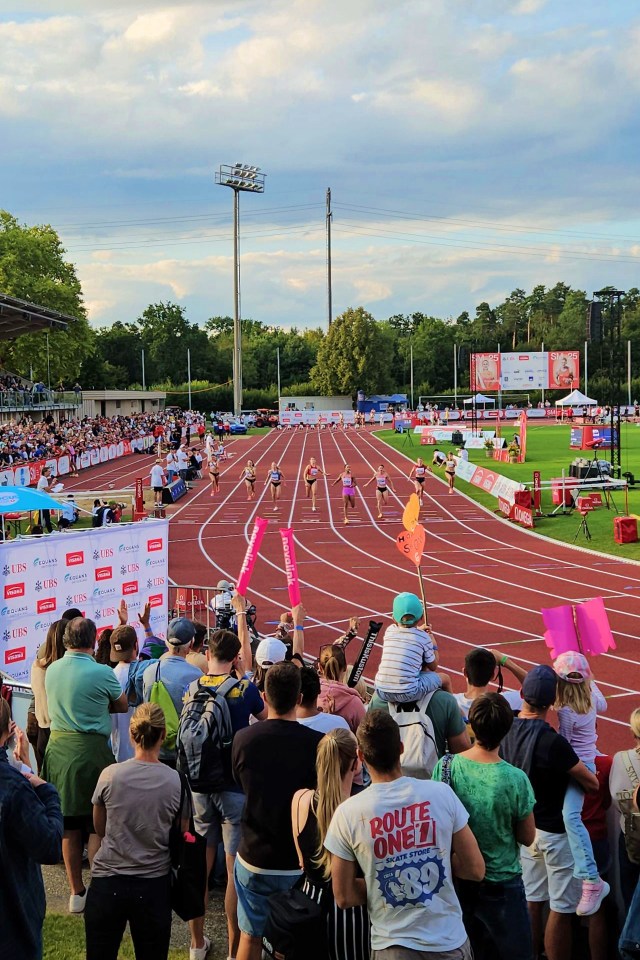 Eine Menschenmenge verfolgt an einem sonnigen Tag ein Sprintrennen der Frauen in einem Leichtathletikstadion im Freien. Die Läuferinnen sind mitten im Rennen, und die Zuschauer halten Schilder und jubeln von der Tribüne und vom Rand der Strecke aus. Im Hintergrund sind Bäume und blauer Himmel zu sehen.