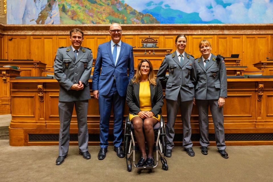 Fünf Personen posieren und lächeln in einem holzgetäfelten Raum; vier tragen Militäruniformen und ein Mann in einem blauen Anzug steht neben einer Frau in einem gelben Kleid, die in einem Rollstuhl sitzt. Über ihnen ist eine bunte Wandmalerei zu sehen.