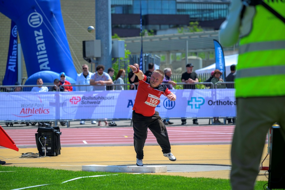 Ein männlicher Athlet in rotem Shirt und schwarzer Hose wirft bei einer Leichtathletik-Veranstaltung Kugelstoßen. Zuschauer beobachten den Wettkampf hinter einer mit Bannern geschmückten Absperrung. Im Vordergrund steht ein Schiedsrichter in einer gelben Weste.