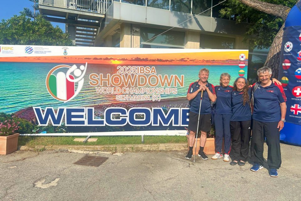 Vier Personen in passender Sportkleidung stehen lächelnd vor einem großen Banner mit der Aufschrift "2025 IBSA Showdown World Championships, Silvi Marina (IT), WELCOME" im Freien, mit Flaggen und einem Sonnenaufgang im Hintergrund.