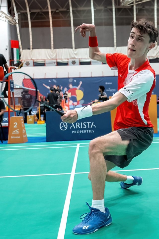 Ein Badmintonspieler in einem rot-weißen Trikot stürmt auf dem Spielfeld nach vorne und bereitet sich auf einen flachen Schlag vor. Sein Gesichtsausdruck ist konzentriert, im Hintergrund sind andere Spieler und Zuschauer zu sehen.