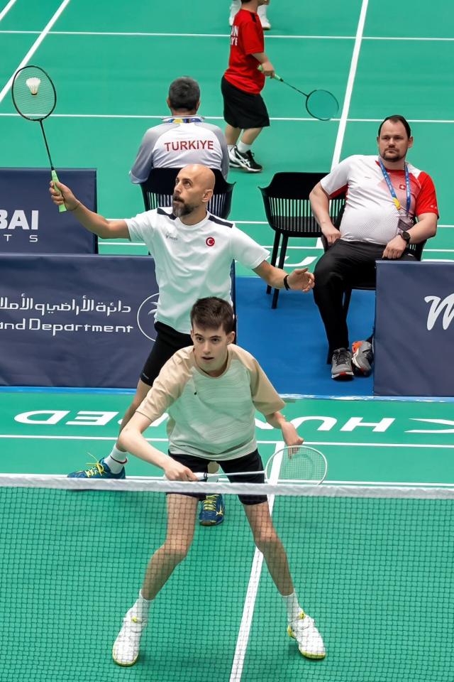 Zwei türkische Badmintonspieler stehen auf einem grünen Hallenplatz bereit, einer am Netz, einer dahinter, und bereiten sich auf einen Return vor. Ein Schiedsrichter sitzt im Hintergrund und beobachtet das Spiel.