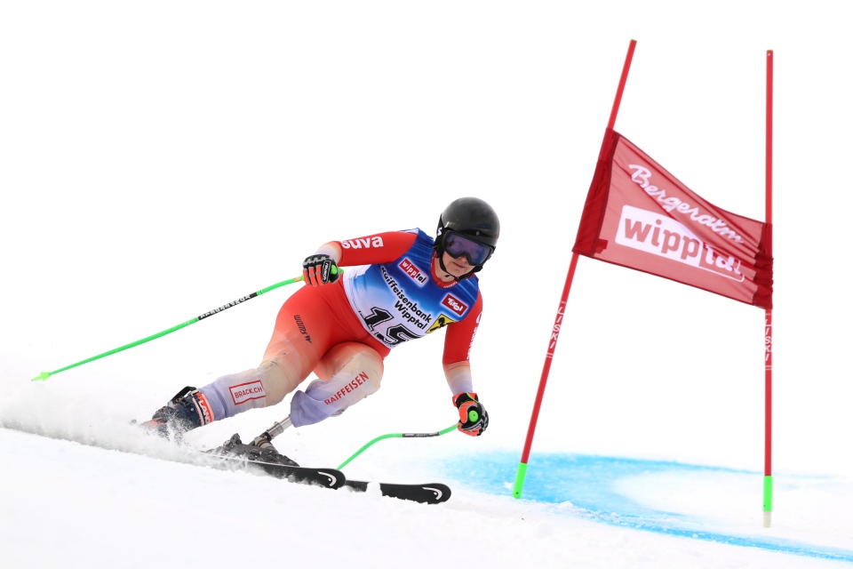 Ein Skifahrer im rot-weißen Rennanzug und mit schwarzem Helm macht während eines alpinen Skirennens eine scharfe Kurve um eine rote Torflagge auf einer verschneiten Piste.