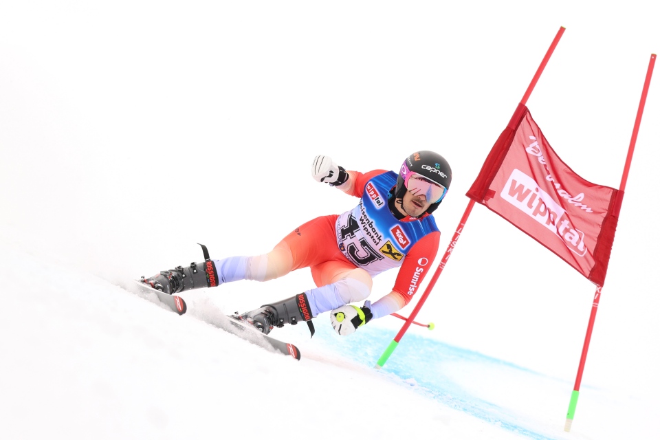 Ein Skifahrer in rot-blauer Rennkleidung und mit schwarzem Helm macht während eines Skirennens auf einer verschneiten Piste eine scharfe Kurve um ein rotes Tor.