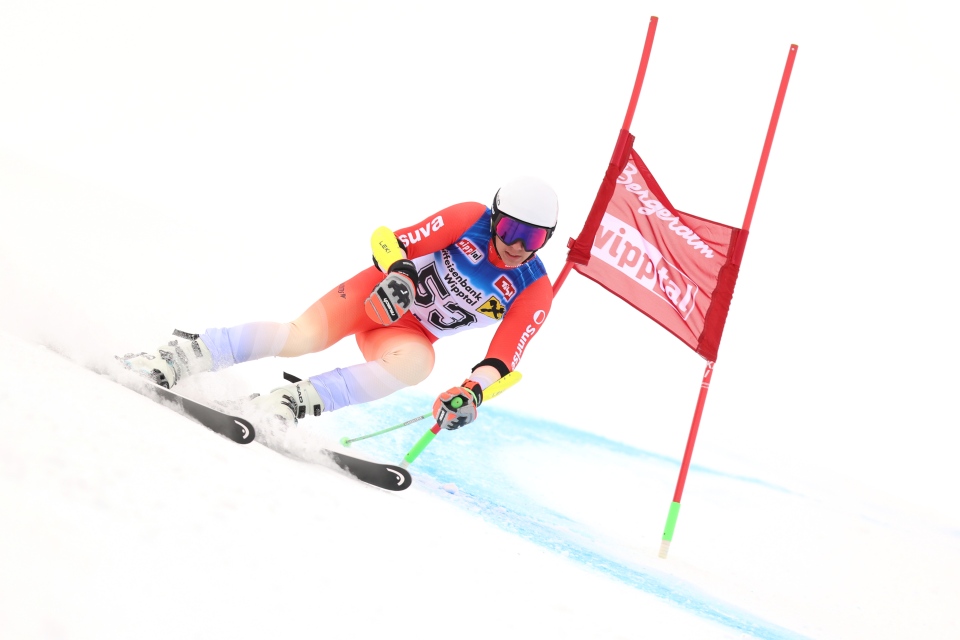 Ein Skifahrer in einem rot-weißen Anzug und Helm fährt während eines Skirennens um ein rotes Slalomtor auf einer verschneiten Piste. Der Skifahrer ist stark gebeugt, mit ausgefahrenen Stöcken und konzentrierter Körperhaltung.