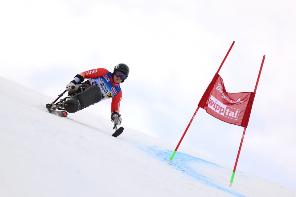 Ein alpiner Skifahrer mit Helm und roter Startnummer fährt während eines Wettkampfs auf einer verschneiten Piste an einer roten Torflagge vorbei bergab.