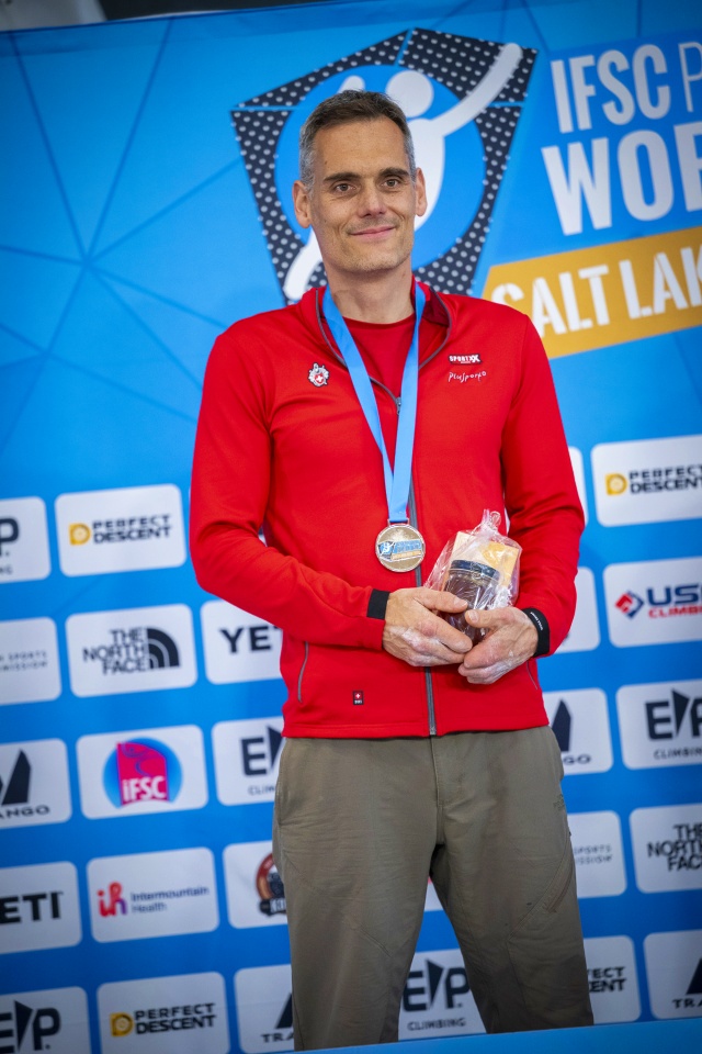 Ein lächelnder Mann in roter Jacke und mit Medaille steht vor einem Banner des IFSC Paraclimbing World Cup Salt Lake City und hält einen kleinen Preis in der Hand. Im Hintergrund sind Sponsorenlogos zu sehen.