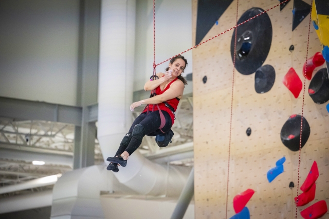 Eine Frau in Sportkleidung hängt beim Klettern in der Halle an einem Seil, lächelt und winkt mit einer Hand in der Nähe einer hohen Kletterwand mit bunten Griffen.