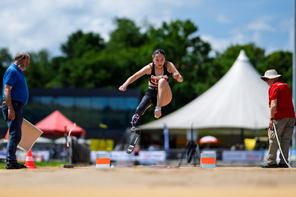 Eine Sportlerin mit einer Beinprothese schwebt beim Weitsprung bei einem Outdoor-Leichtathletik-Event in der Luft, zwei Offizielle schauen zu, im Hintergrund sind Zelte zu sehen.
