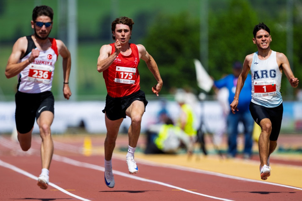 Drei männliche Athleten sprinten während eines Rennens auf einer Freiluftbahn. Jeder trägt Startnummern und Sportkleidung. Im Hintergrund sind verschwommenes Grün und Zuschauer zu sehen.