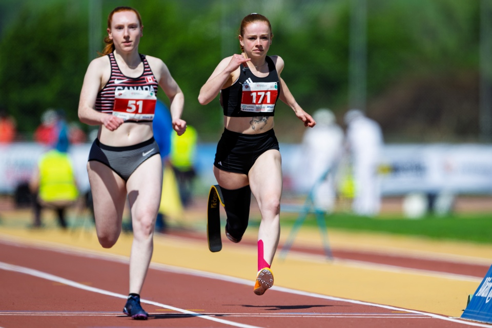 Zwei Sportlerinnen sprinten konzentriert und mitten im Schritt auf einer Laufbahn. Die vordere Läuferin trägt die Nummer 171 und einen rosa Kompressionsstrumpf, die andere, in einem gestreiften Oberteil, die Nummer 51.