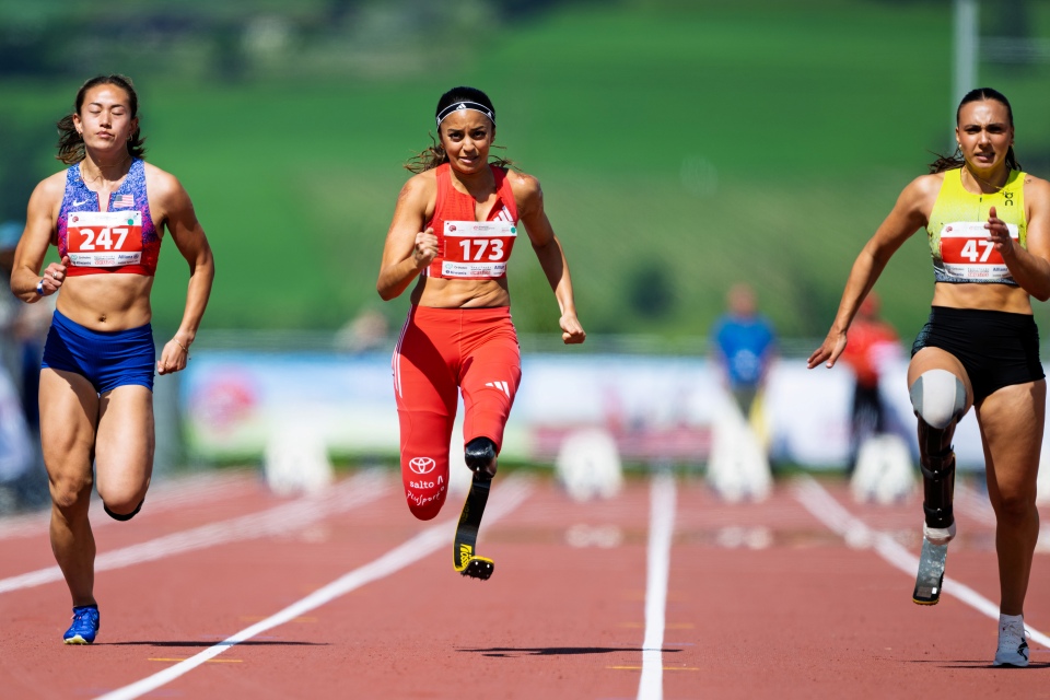 Drei Para-Athletinnen sprinten auf einer Laufbahn. Die Läuferinnen in der Mitte und rechts tragen Beinprothesen. Sie tragen Startnummern und blicken entschlossen. Im Hintergrund ist ein verschwommenes grünes Feld zu sehen.
