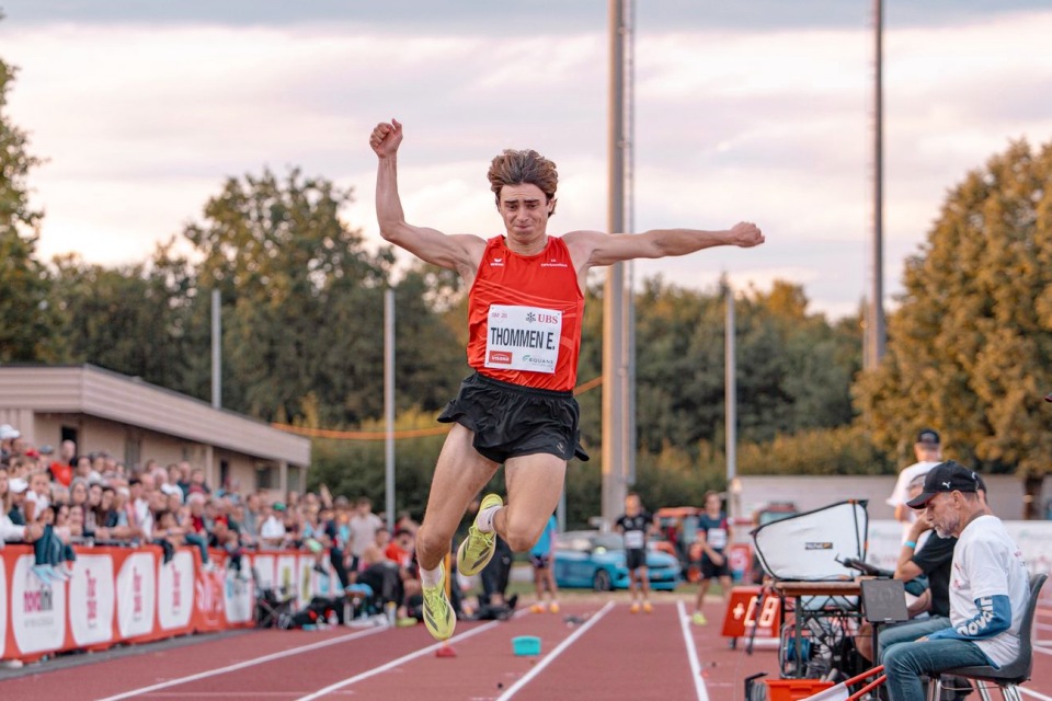 Ein männlicher Athlet in einem roten Trikot und schwarzen Shorts befindet sich während eines Weitsprungs bei einem Leichtathletik-Wettkampf im Freien in der Luft, während im Hintergrund Zuschauer und Offizielle zusehen.