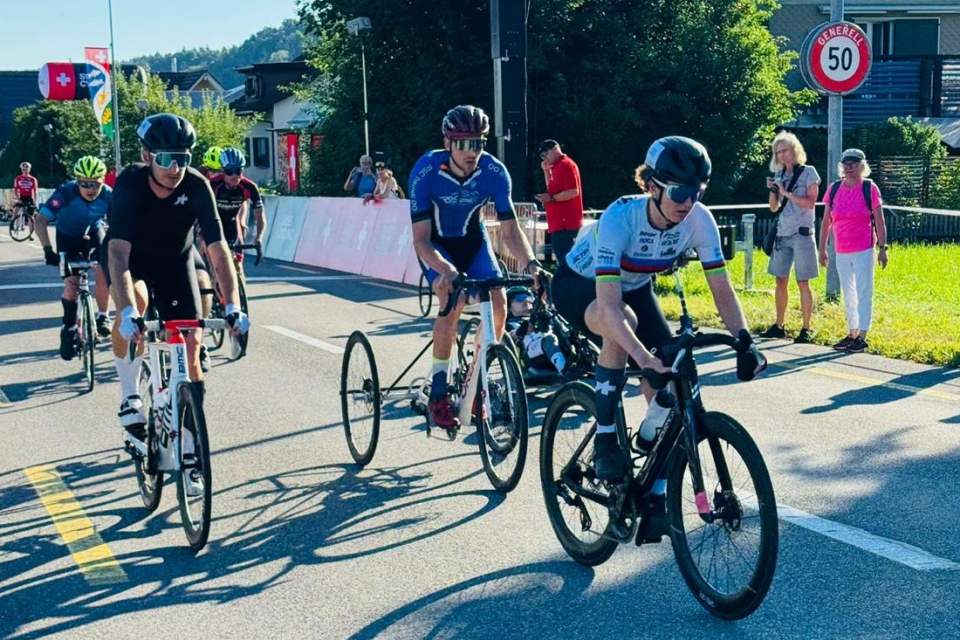 Radfahrer nehmen an einem sonnigen Tag an einem Straßenrennen teil und fahren entlang einer von Zuschauern gesäumten Straße. Im Hintergrund sind zwischen Häusern und Grünflächen ein Schild mit einer Geschwindigkeitsbegrenzung von 50 km/h und die Schweizer Flagge zu sehen.