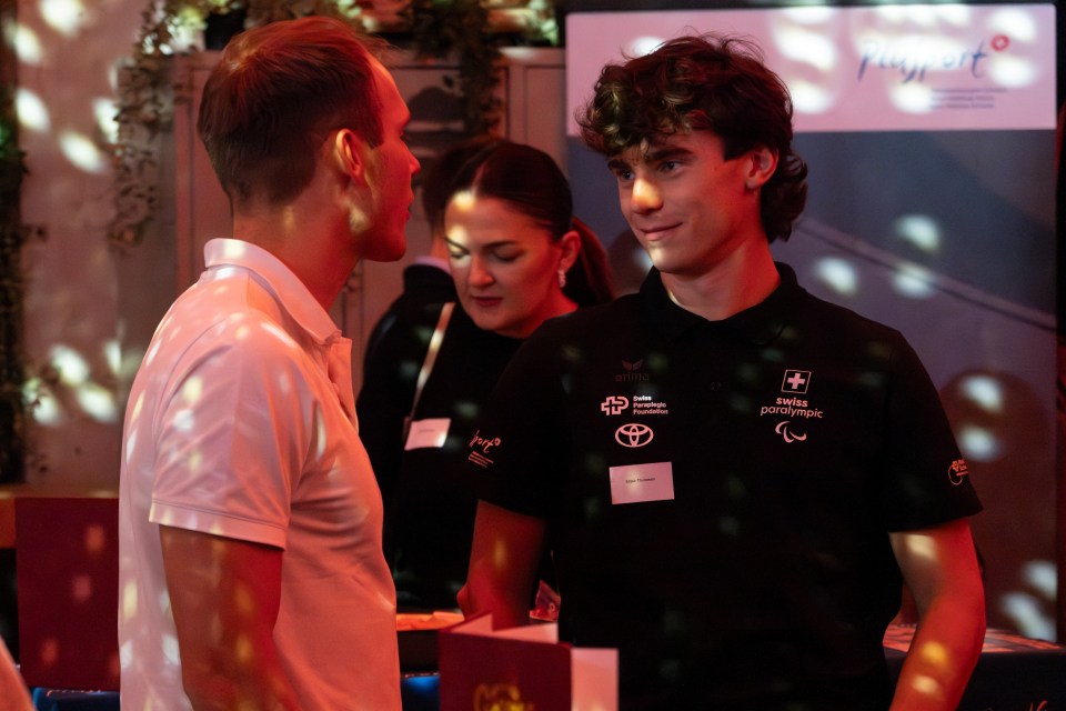 Zwei junge Männer stehen und unterhalten sich in einem warm beleuchteten Innenraum mit festlichen Lichtmustern; eine Frau arbeitet an einem Tisch im Hintergrund. Der Mann auf der rechten Seite trägt ein schwarzes Polohemd mit Logos.