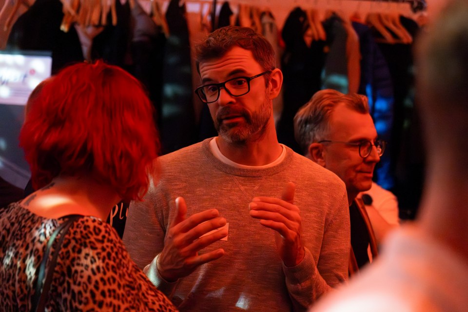 Ein Mann mit Brille und grauem Pullover gestikuliert, während er sich mit einer Frau mit kurzen roten Haaren und einem Oberteil mit Leopardenmuster bei einer gesellschaftlichen Veranstaltung in einem Gebäude unterhält. Ein weiterer Mann ist im Hintergrund zu sehen.