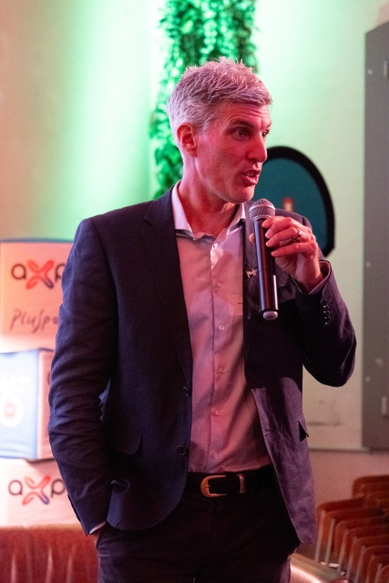 Ein Mann in einem dunklen Anzug hält ein Mikrofon in der Hand und spricht bei einer Indoor-Veranstaltung, mit grüner Beleuchtung und Markenboxen im Hintergrund.