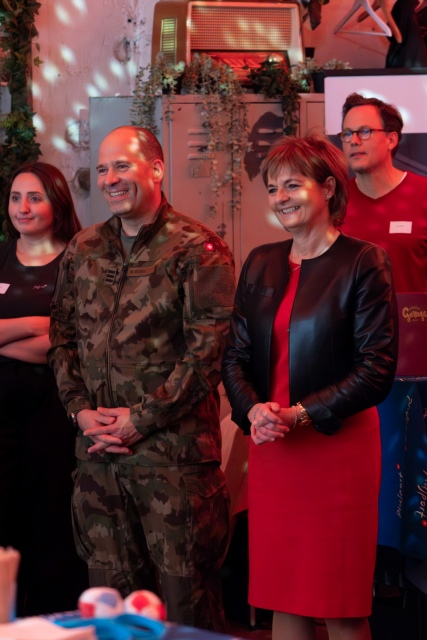 Eine Gruppe von Menschen steht lächelnd in einem Haus. Ein Mann trägt eine Militäruniform in Tarnfarben, während eine Frau neben ihm ein rotes Kleid und eine schwarze Jacke trägt. Andere leger gekleidete Menschen stehen im Hintergrund inmitten von dekorativen Lichtern.