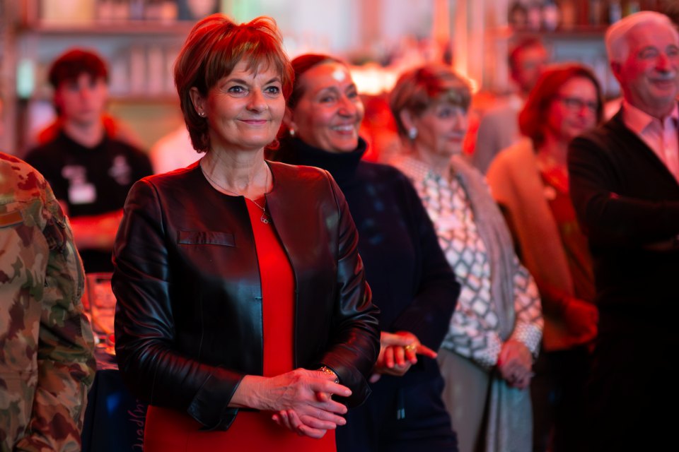 Eine Frau in einem roten Kleid und einer schwarzen Lederjacke steht lächelnd in einem warm beleuchteten Raum, umgeben von anderen Menschen, die ebenfalls lächeln und nach vorne schauen.