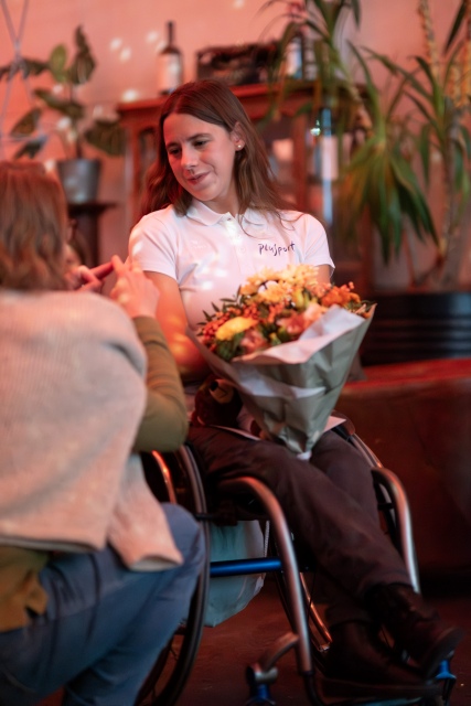 Eine Frau im Rollstuhl hält einen Blumenstrauß und lächelt eine andere Person an. Sie trägt ein weißes Polohemd, und im Hintergrund sind Pflanzen und Flaschen zu sehen.