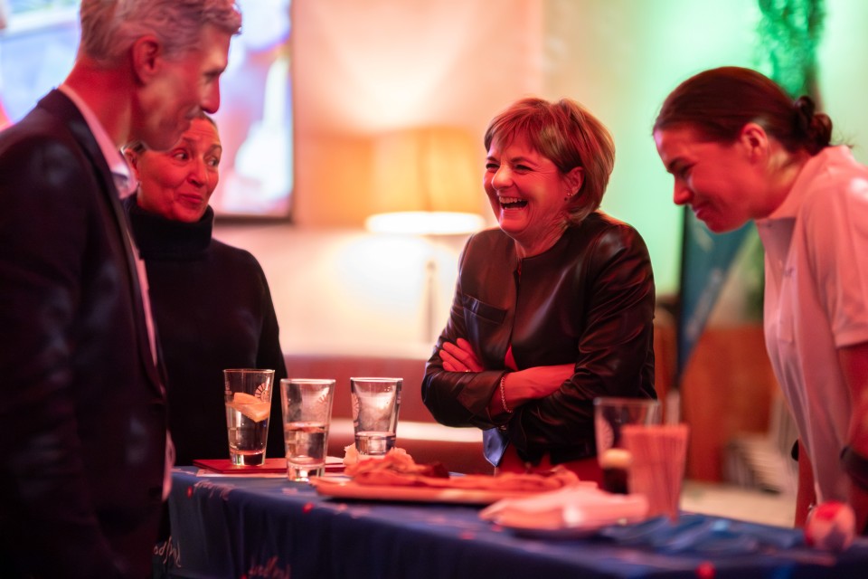 Vier Personen stehen um einen hohen Tisch mit Getränken und Snacks und unterhalten sich angeregt. Eine Frau in einer schwarzen Jacke lacht, während die anderen lächeln und zuhören. Der Hintergrund ist sanft beleuchtet und bunt.