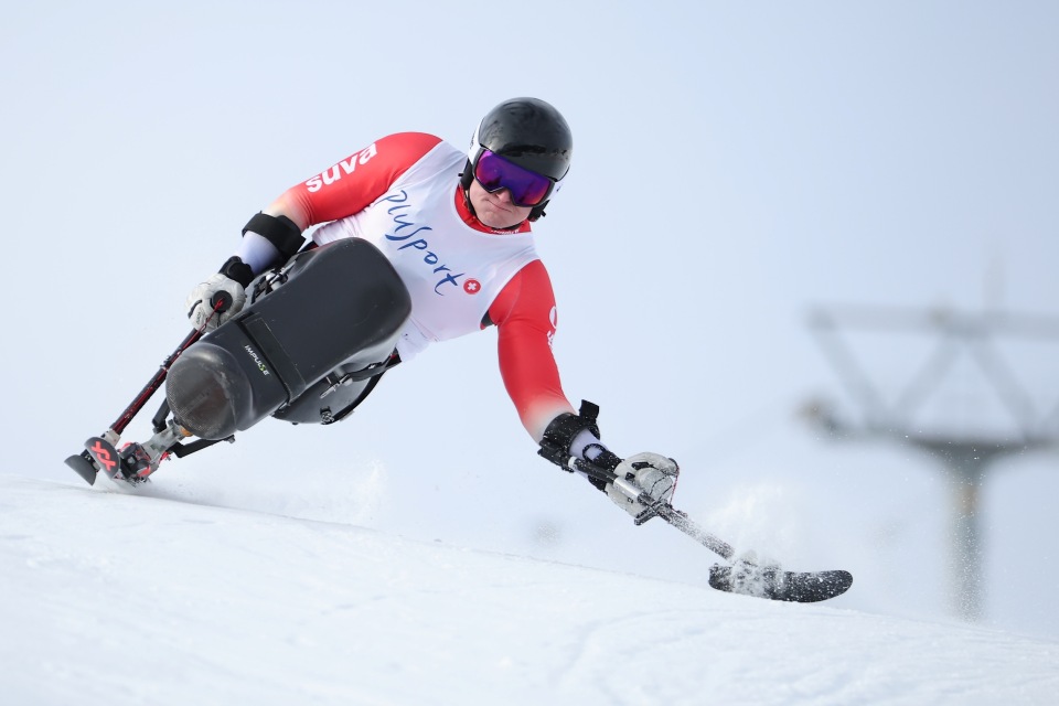 Ein Alpinskifahrer in rot-weißer Ausrüstung fährt mit einem Sitzski und Auslegerstöcken eine scharfe Kurve auf einer verschneiten Piste. Der Schnee spritzt von den Skiern, und im Hintergrund ist ein Skilift zu erkennen.
