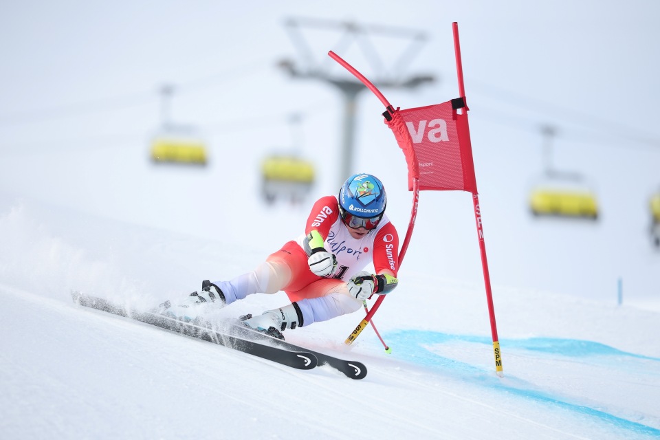 Ein Skifahrer in einem weiß-roten Anzug und mit Helm fährt bergab und neigt sich stark, während er ein rotes Slalomtor durchfährt. Im verschwommenen Hintergrund sind Schneespritzer und Skilifte zu sehen.