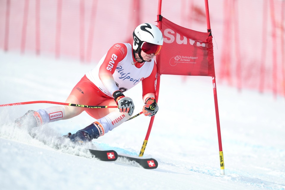 Ein Skifahrer in einem rot-weißen Anzug und mit Helm macht eine scharfe Kurve um ein rotes Slalomtor auf einer verschneiten Piste während eines Skirennens, mit rotem Netz im Hintergrund.