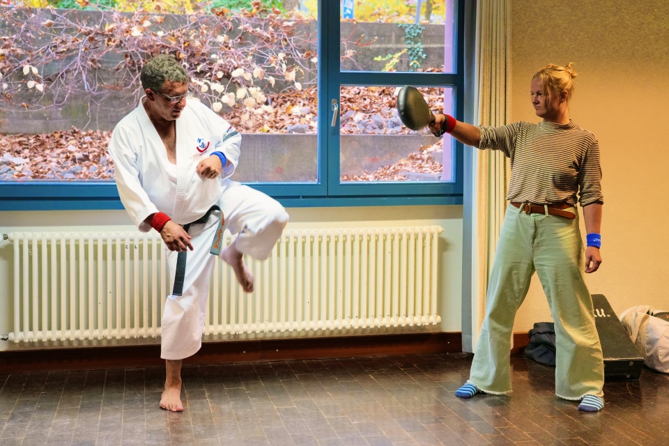 Ein älterer Mann in einem weißen Kampfsportanzug übt in einer Halle einen hohen Tritt und zielt auf ein gepolstertes Ziel, das von einer jüngeren Frau in Freizeitkleidung gehalten wird, die in der Nähe eines Fensters steht.