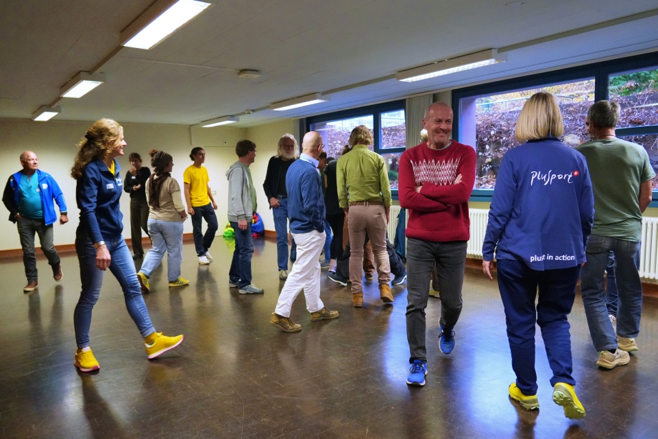 Eine Gruppe Erwachsener geht in einem großen, hell erleuchteten Raum mit großen Fenstern umher. Die Menschen sind leger gekleidet und scheinen an einer Indoor-Aktivität oder einem Workshop teilzunehmen.