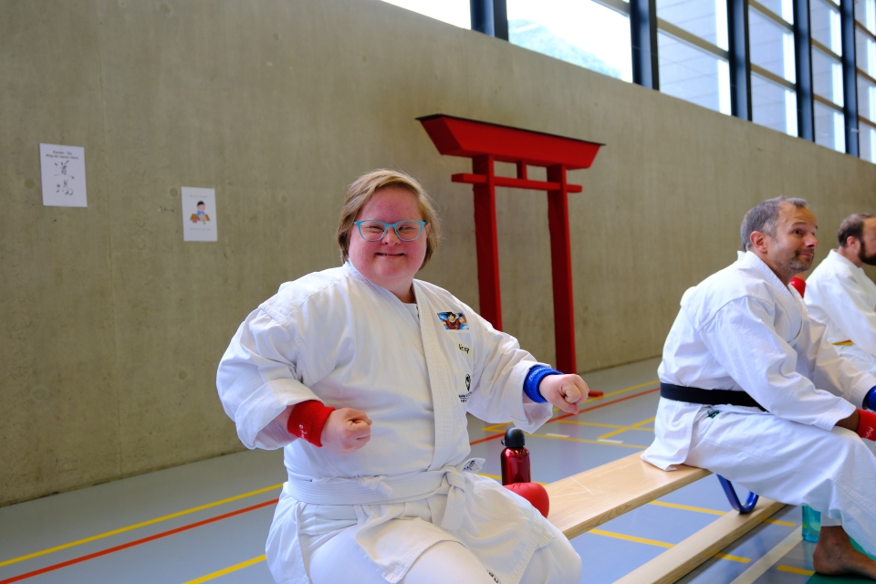 Eine lächelnde Person in einem Karateanzug sitzt auf einer Bank in einer Turnhalle und macht eine Pose mit zwei Fäusten. Zwei weitere Personen in Karateuniformen sitzen in der Nähe. Im Hintergrund sind ein rotes Torii-Tor und Plakate zu sehen.