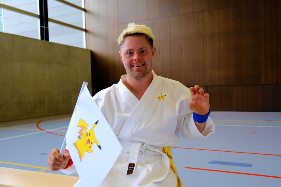 Ein lächelnder Mann in einem Karateanzug hält eine Fahne mit einer Pikachu-Grafik in einer Sporthalle. Er trägt einen gelben Gürtel, ein blaues Armband und gestikuliert mit der freien Hand. Die Turnhalle hat Wände aus Holz und Beton.