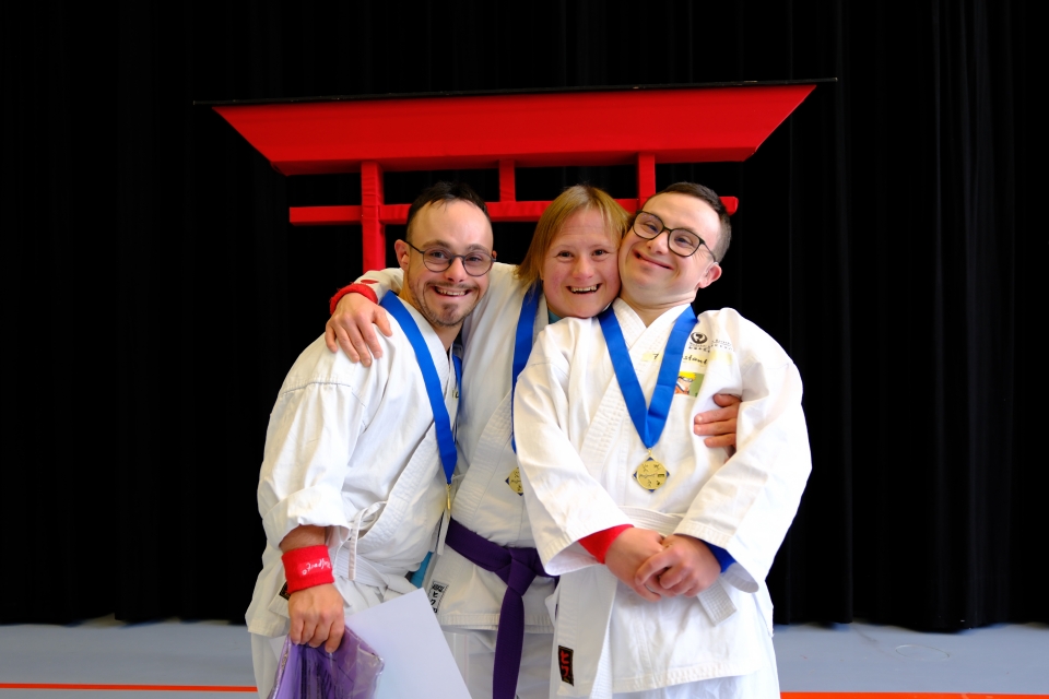 Drei Personen, die Kampfsportuniformen und Medaillen tragen, stehen lächelnd und sich umarmend vor einem roten Gebäude mit dunklem Hintergrund.