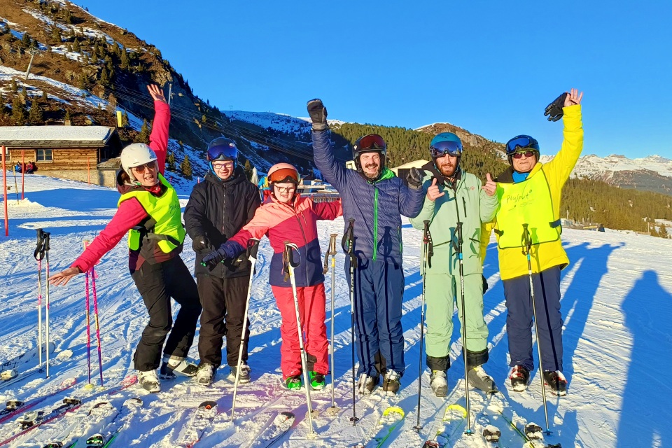 Sechs Personen in farbenfroher Skikleidung stehen auf einer verschneiten Piste, lächeln und posieren mit erhobenen Armen und Skistöcken, mit Bergen, Bäumen und einer Holzhütte im Hintergrund unter einem klaren blauen Himmel.