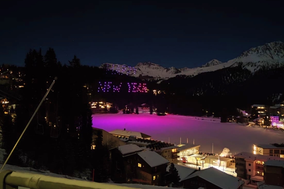 Ein verschneites Bergdorf bei Nacht mit dem Schriftzug NEW YEAR in violetten und rosafarbenen Lichtern auf einem Berghang; der gefrorene See darunter ist mit lebhaftem violettem Licht beleuchtet. Im Hintergrund sind schneebedeckte Gipfel zu sehen.