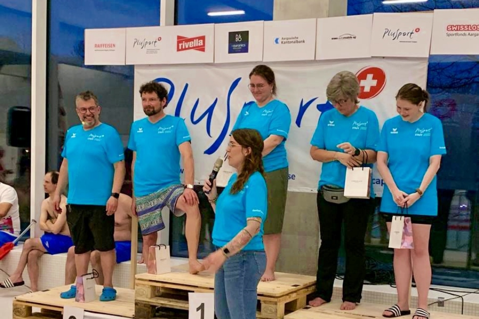 Sechs Personen in blauen Hemden stehen lächelnd auf einem aus Holzpaletten gebauten Siegerpodest bei einem Hallenschwimmfest. Eine Frau hält ein Mikrofon und scheint zu sprechen. Hinter ihnen ist ein Banner mit Logos zu sehen.