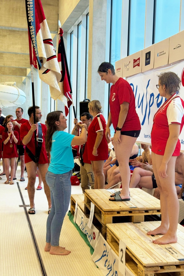 Eine Frau in einem türkisfarbenen Hemd überreicht Medaillen an Sportler in roten Uniformen, die bei einem Hallenschwimmwettbewerb auf einem Podium stehen. Im Hintergrund werden Fahnen gehalten, und Zuschauer schauen von der Seite zu.