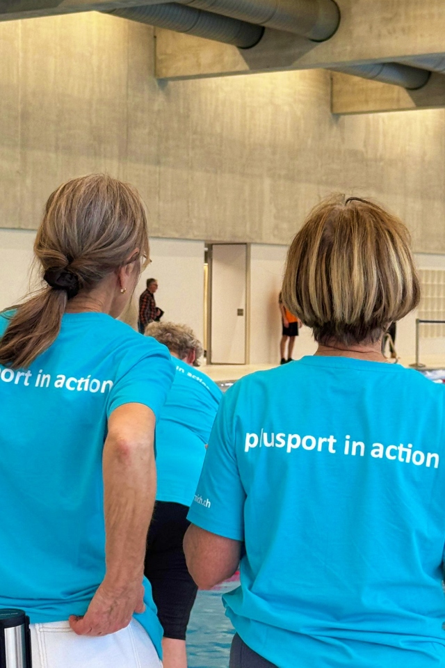 Zwei Frauen in passenden türkisfarbenen plusport in action-T-Shirts stehen an einem Hallenbad und schauen von der Kamera weg. Im Hintergrund sind andere Menschen und Schwimmer zu sehen.