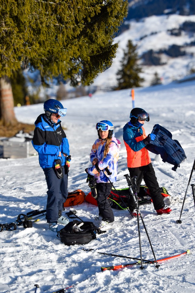 Drei Personen in Skiausrüstung stehen auf einer verschneiten Piste mit Skistöcken und Ausrüstung um sie herum. Einer passt eine Jacke an, ein anderer hält einen Mantel, und der dritte steht in der Nähe. Im Hintergrund sind Bäume und Berge zu sehen.