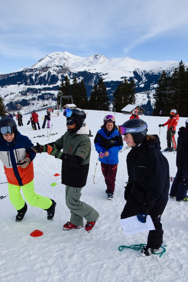 Eine Gruppe von Menschen in Skiausrüstung lacht und unterhält sich auf einer verschneiten Piste mit Bergen und Bäumen im Hintergrund. Einige halten Papiere und Skistöcke, andere scheinen gemeinsam zu spielen oder zu üben.